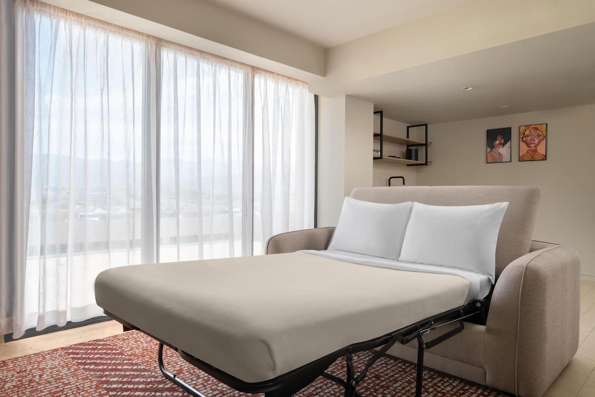 A hotel room with a bed, a window with sheer curtains, a view of the city, and a small shelf with pictures.