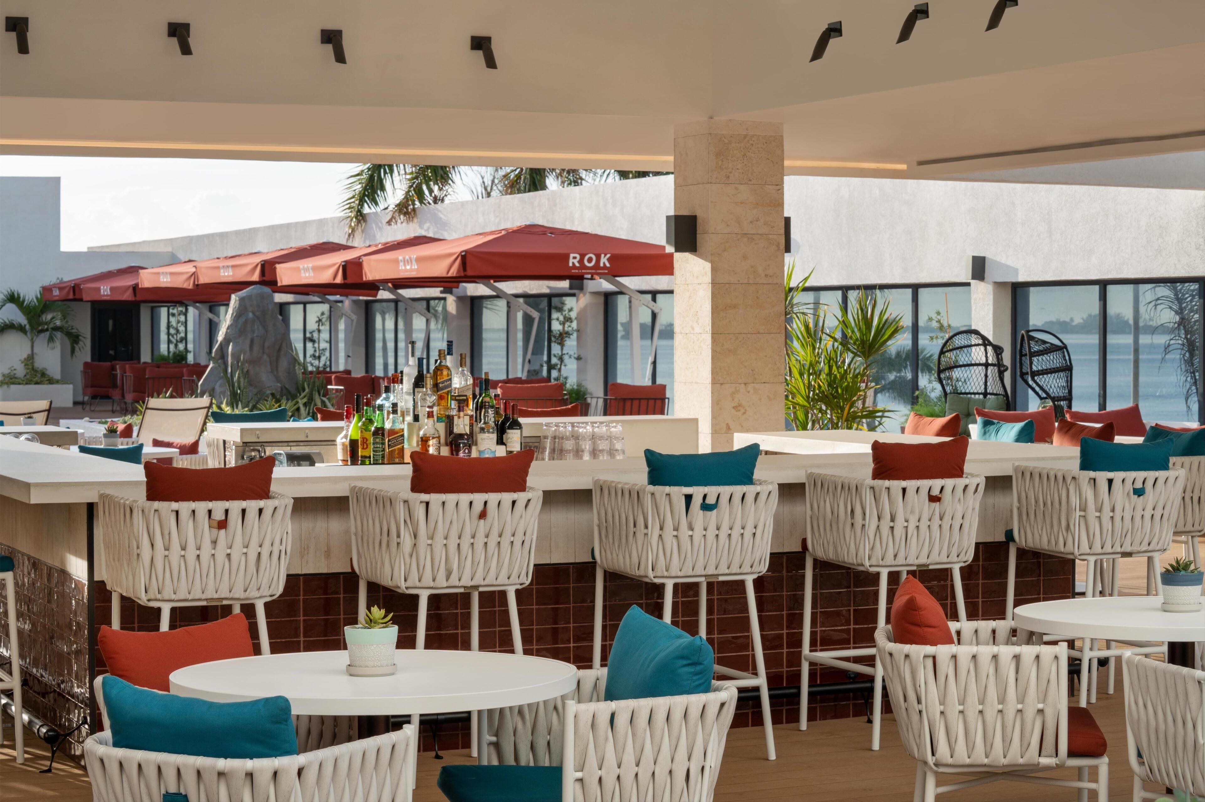 Un espace bar en plein air avec des chaises en osier blanc et des parasols rouges, un comptoir de bar avec des bouteilles, et une vue sur la mer.