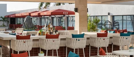Zona de bar al aire libre con sillas de mimbre blanco y sombrillas rojas, una barra con botellas y vistas al mar.
