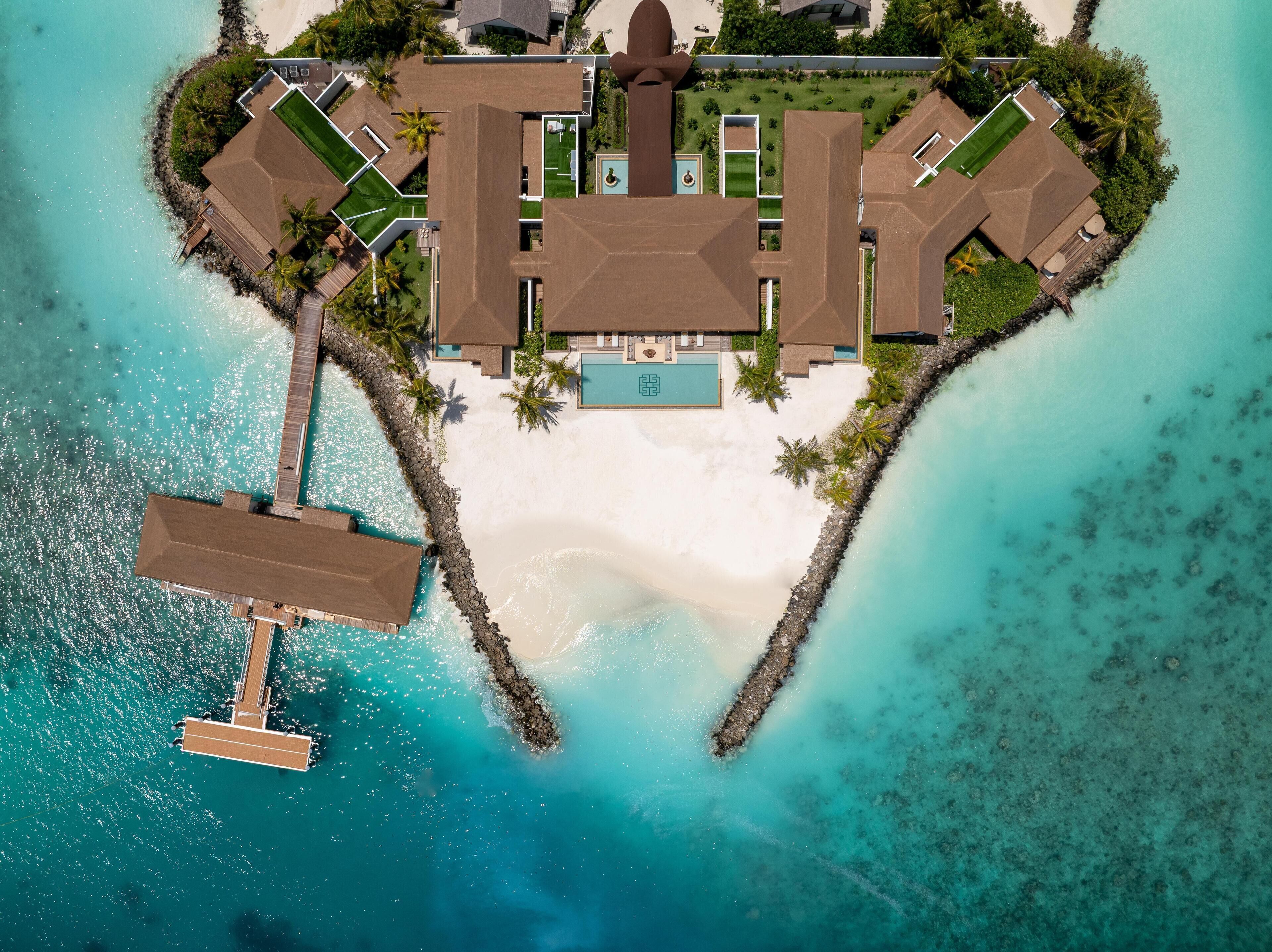 Een luchtbeeld van een resort in een hartvormige indeling, met een centraal zwembad, meerdere gastvillas en een strand.