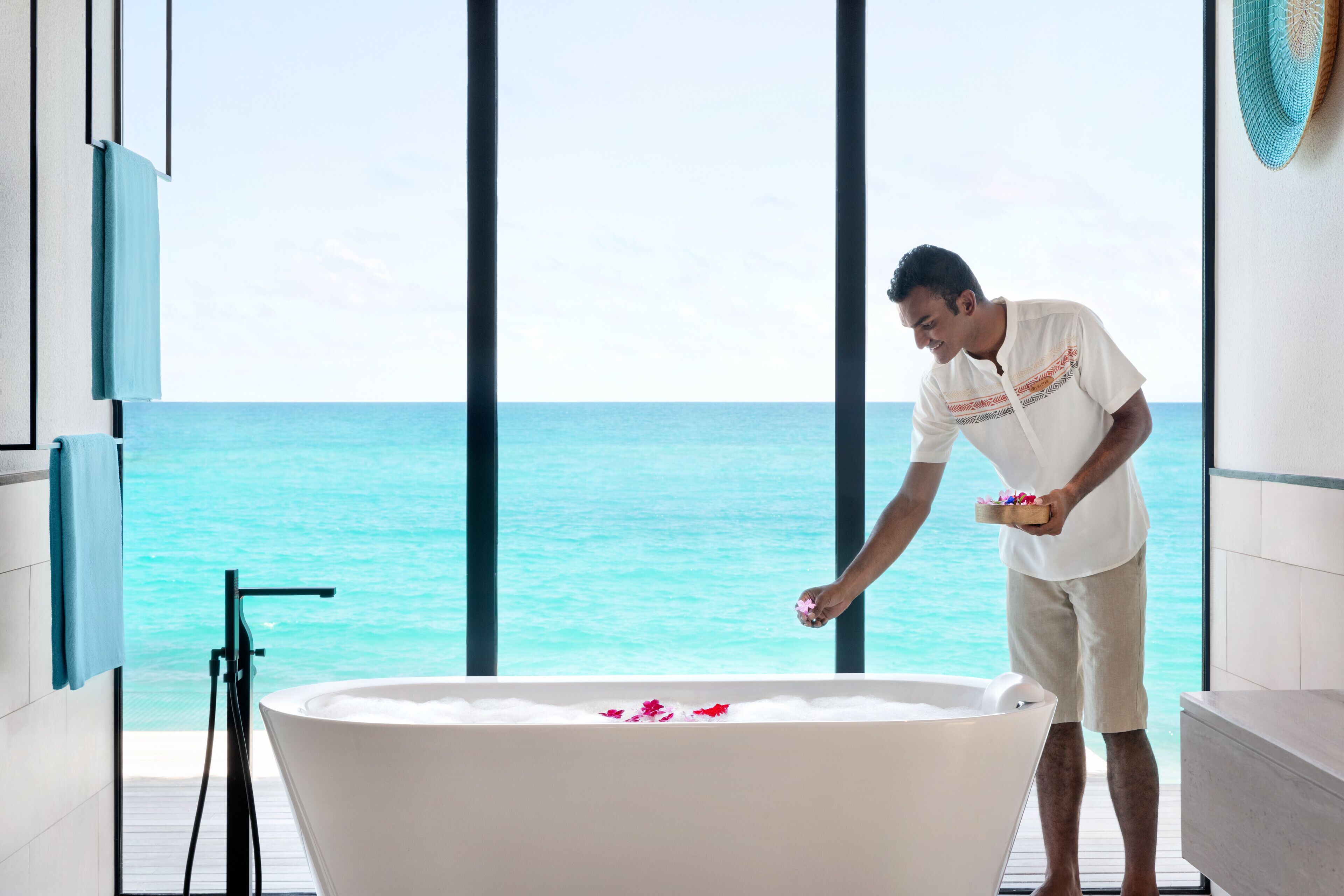 Un homme vêtu d’une chemise blanche et d’un short beige, près d’une baignoire remplie de pétales de rose, avec une vue sur l’océan par de grandes fenêtres.