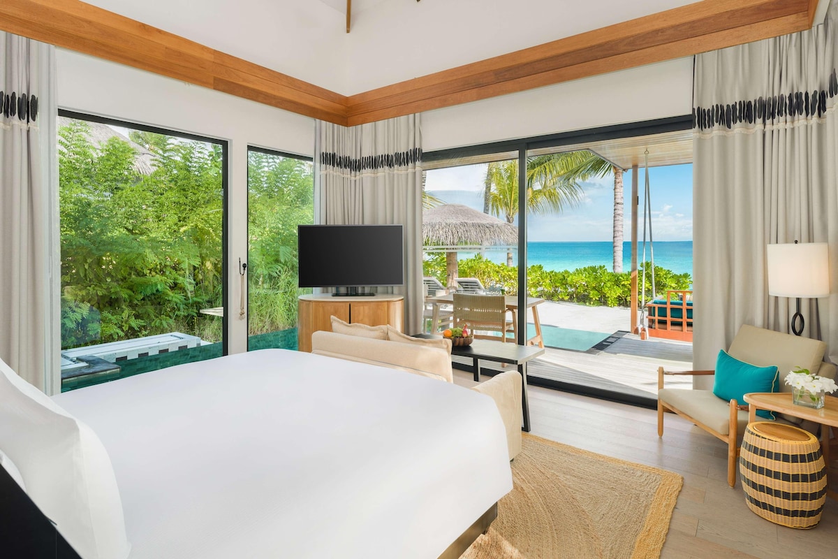 A hotel room with a large bed, a TV, a dining area with a table and chairs, and a view of the ocean.