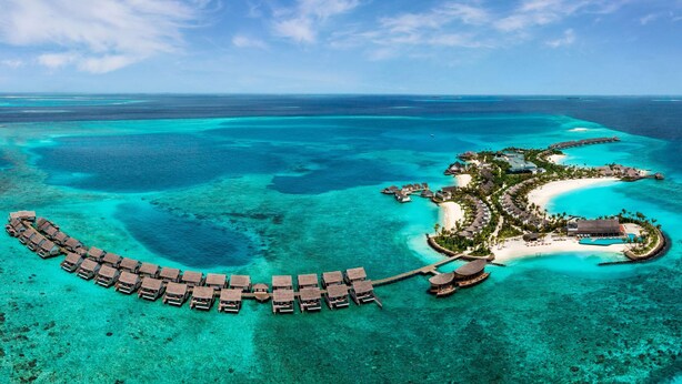 Vue aérienne d’un complexe hôtelier avec des bungalows sur pilotis, une plage et une eau turquoise cristalline.