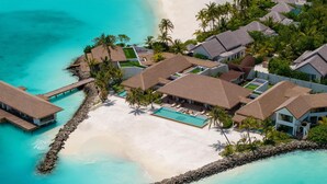 Vue aérienne d’un complexe hôtelier comprenant des bungalows sur pilotis, une piscine centrale et une plage.