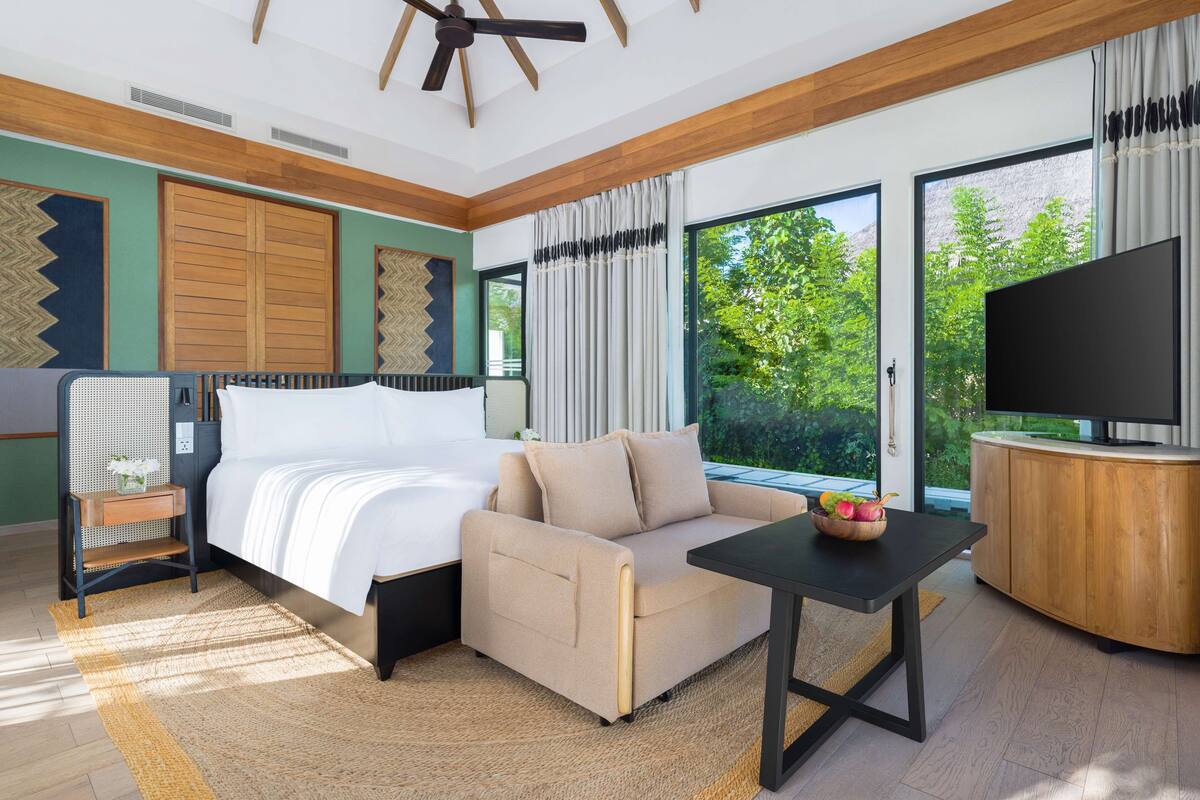 Ein modernes Hotelzimmer mit Bett, Sofa, Fernseher und großen Fenstern mit Blick ins Grüne.