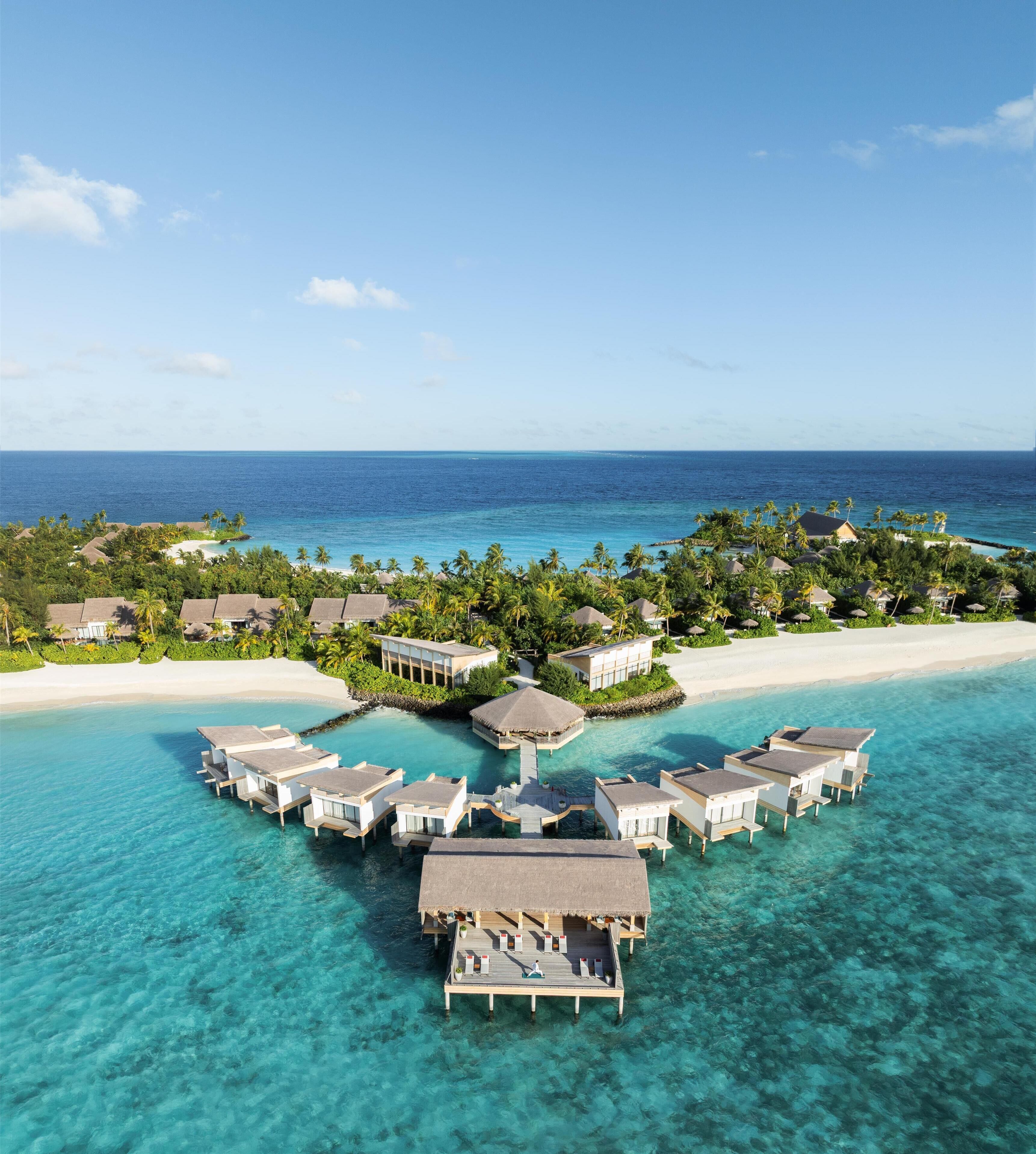 Een overwaterresort met meerdere bungalowachtige accommodaties verbonden door loopbruggen, omgeven door helder turquoise water en een zandstrand.