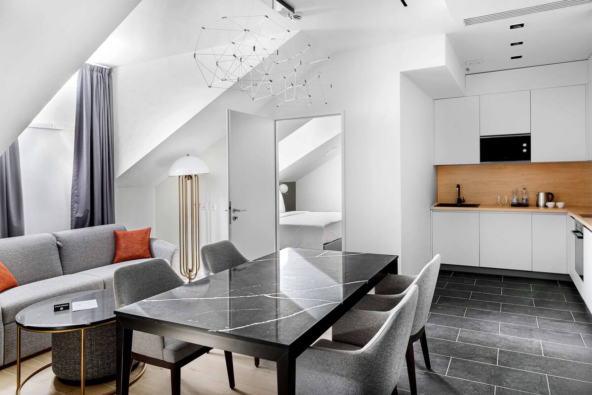 A modern living room with a dining table, grey sofa, and a kitchen area with white cabinets and a black countertop.
