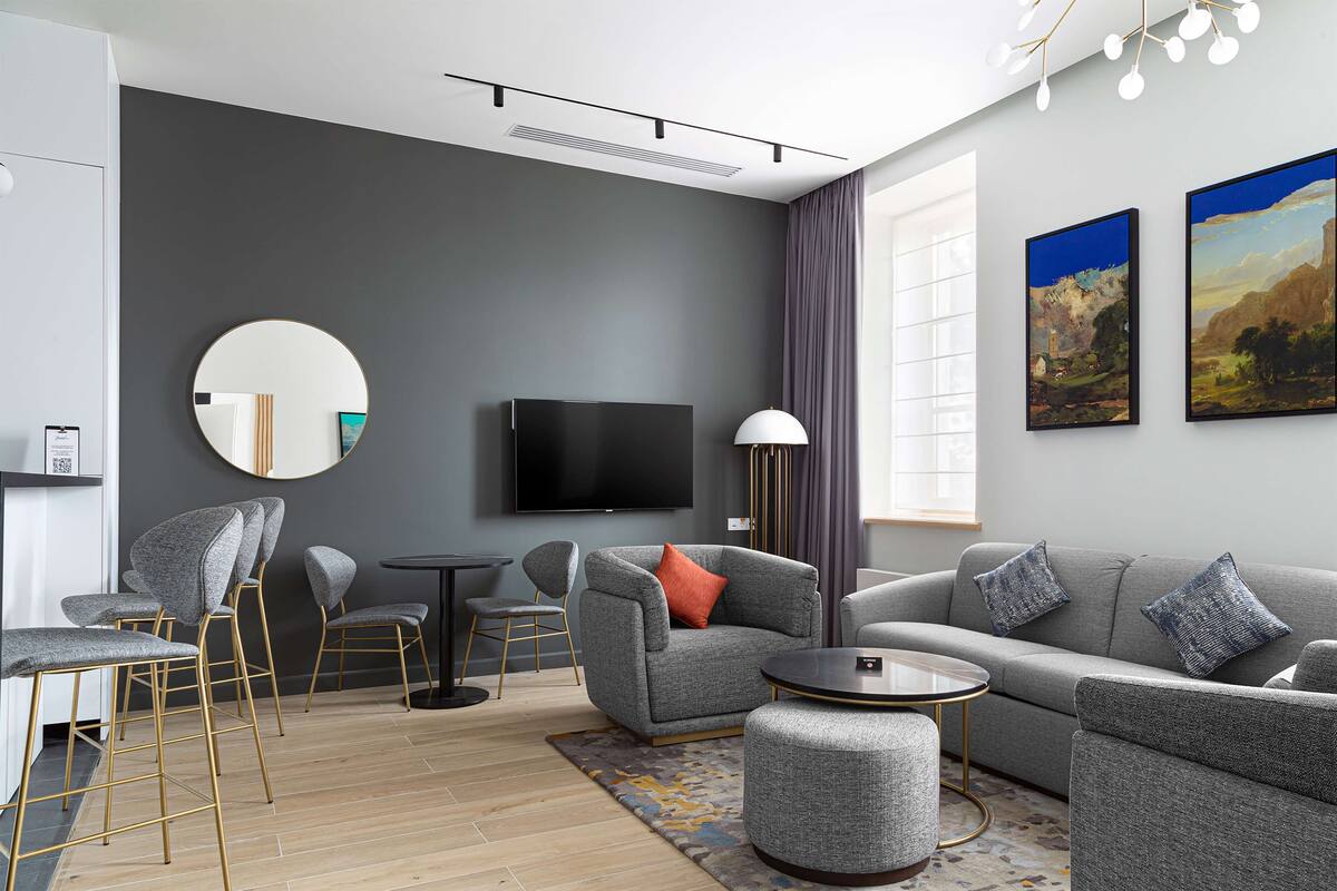 A modern living room with grey sofas, a round mirror, a flat-screen TV, and a dining area with bar stools.
