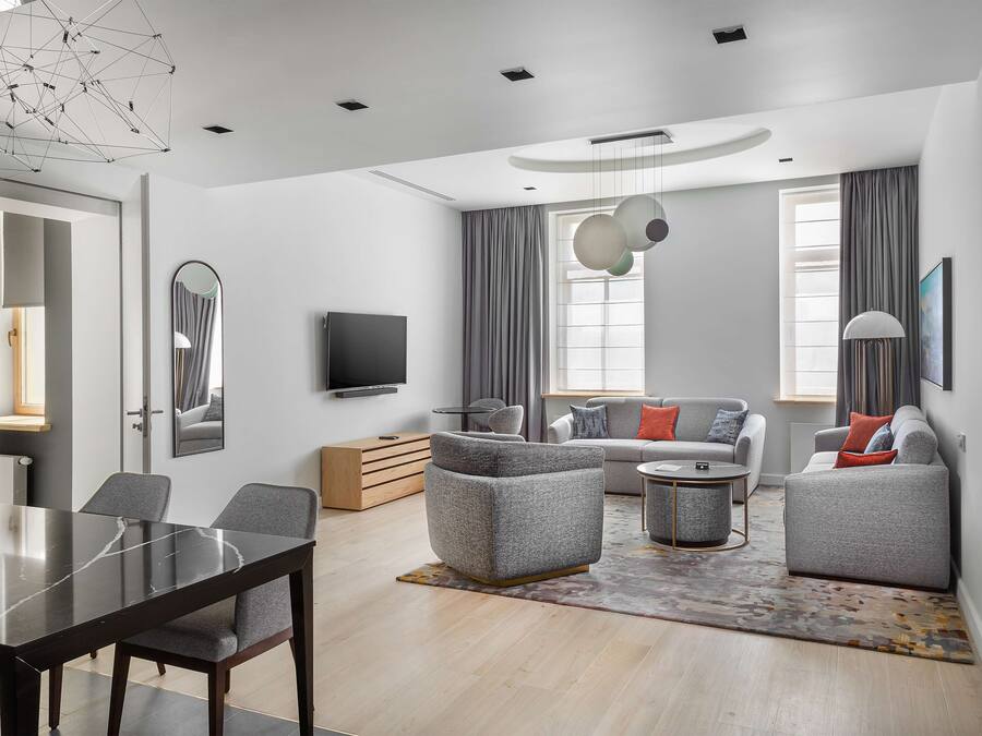 A modern living room with a dining table, grey sofas, a TV, and a large window with curtains.