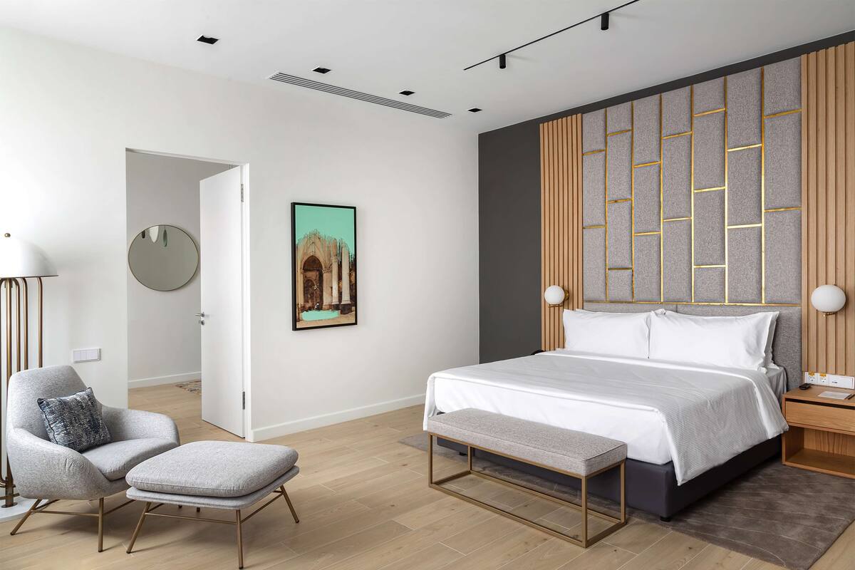 A modern bedroom with a bed, a chair, a bench, a bedside table, a lamp, a wall-mounted mirror, and a framed picture.