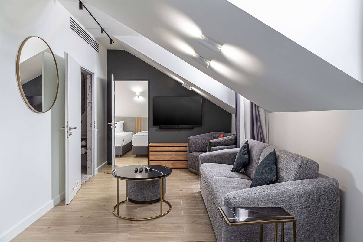A modern living room with a grey sofa, a round mirror, a small round table, and a flat-screen TV mounted on the wall.