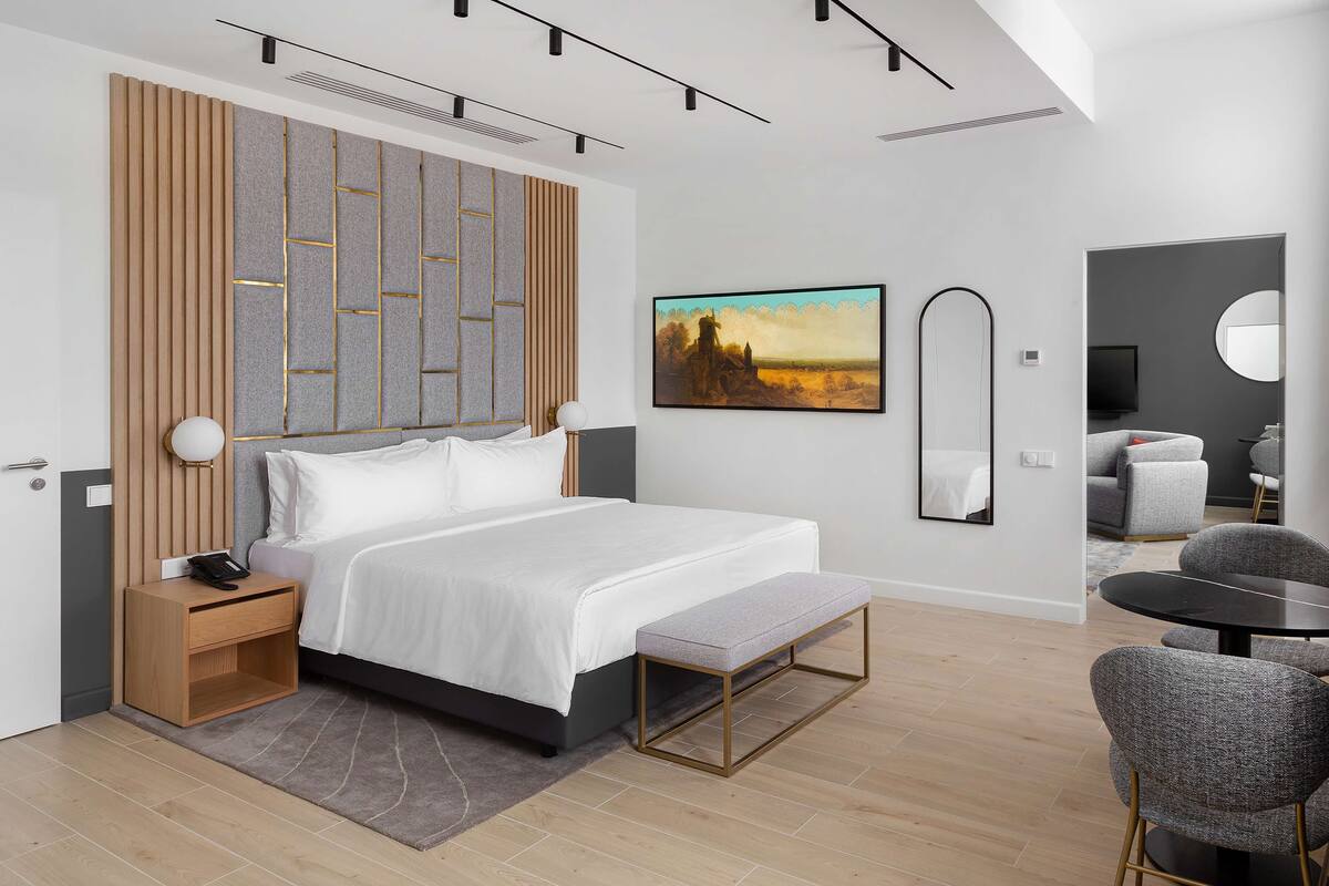 A modern hotel room with a large bed, bedside table, and a small desk. There is a bench at the foot of the bed, a round table with chairs, and a large mirror on the wall.