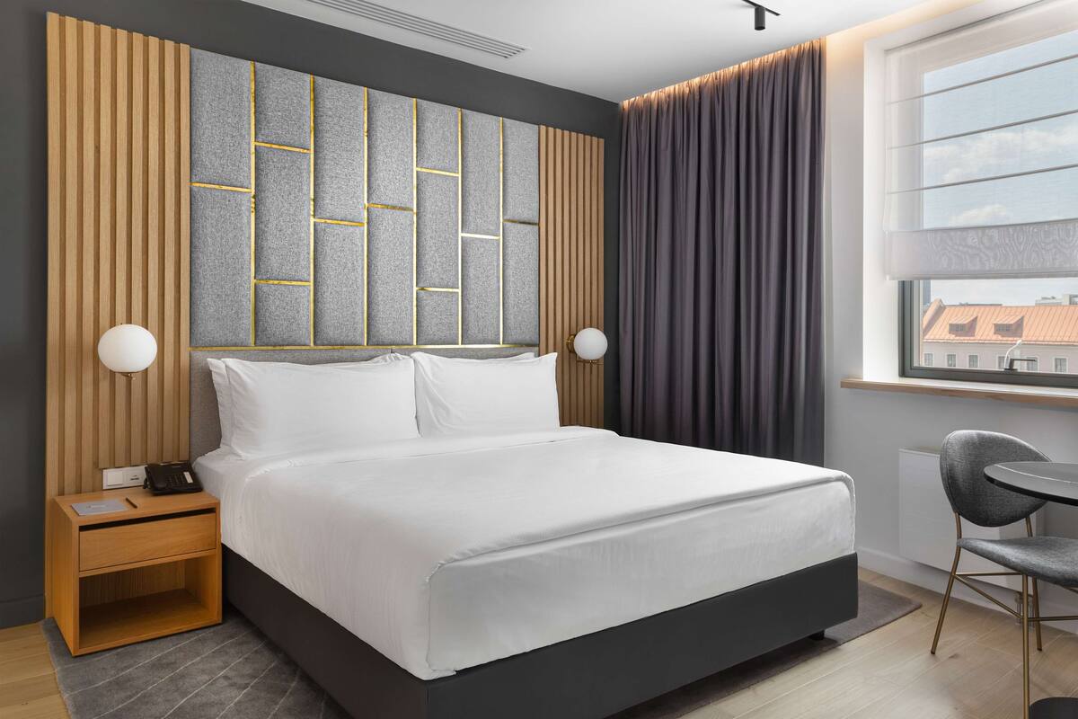 A modern hotel room with a large bed, wooden headboard, and a view of the city through a window.