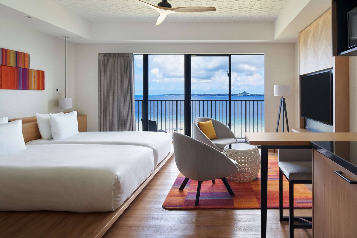 Ein Hotelzimmer mit zwei Betten, einem Fernseher, einem Balkon mit Meerblick und einem Essbereich.