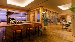 Een moderne bar met een houten toog en oranje barstoelen, grote ramen en uitzicht op de zonsondergang.