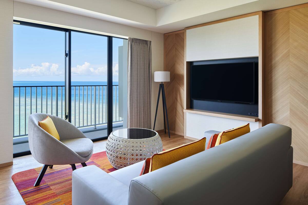Ein modernes Hotelzimmer mit Balkon, einem großen Flachbildfernseher, einem grauen Sessel, einem weißen Sofa und einem gemusterten Hocker.