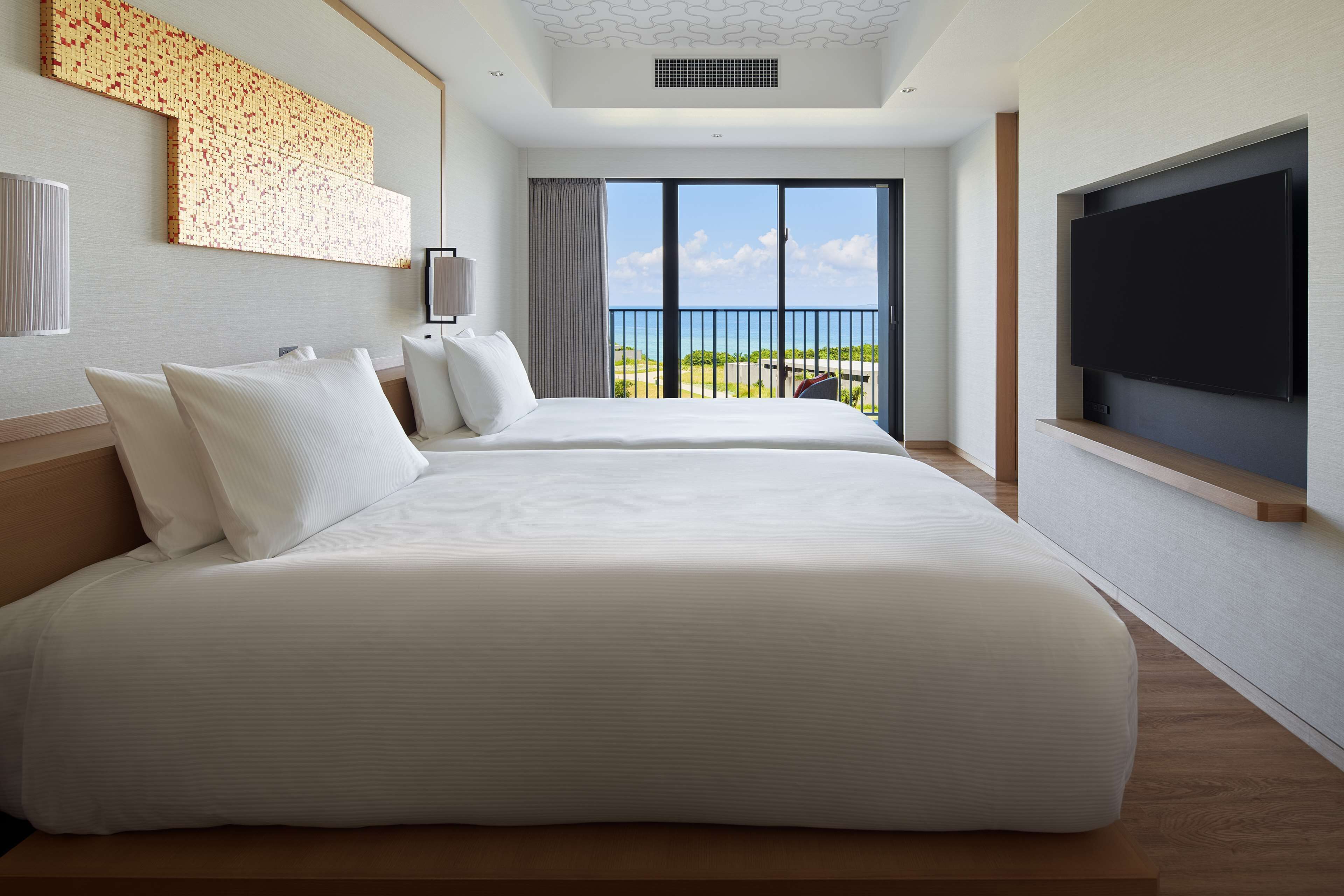 A modern hotel room with two large beds, a flat-screen TV, and a balcony with a view of the sea.
