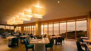 Een ruim restaurant met hoge plafonds, grote ramen met uitzicht op de zee en moderne verlichtingsarmaturen.