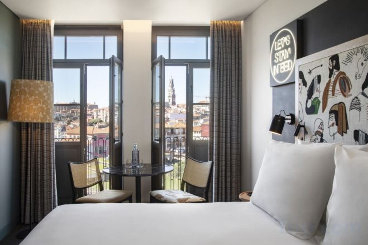 A hotel room with a bed, a small table with chairs, a view of the city through large windows, and a wall with a poster.