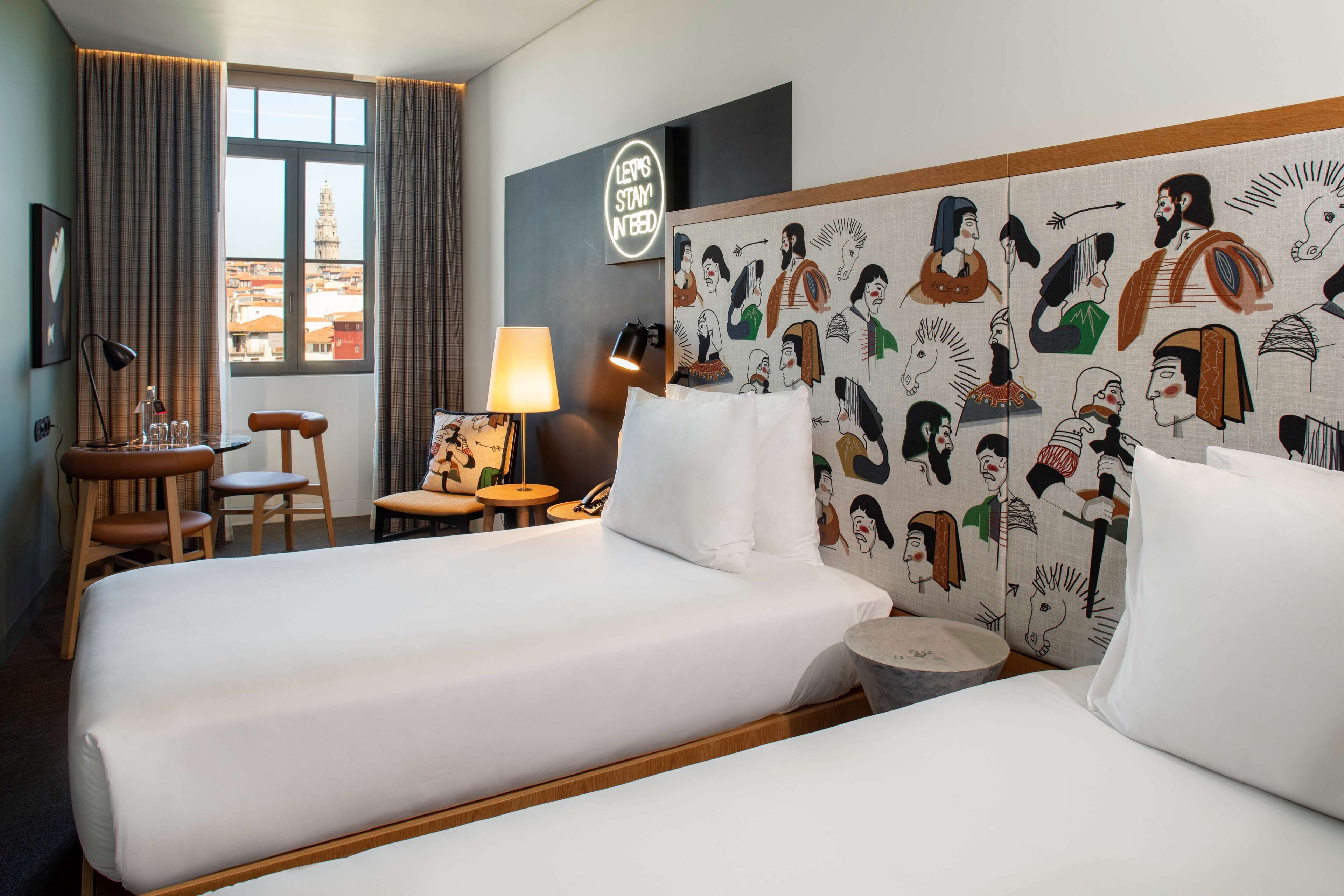 Habitación de hotel con dos camas, un escritorio, sillas, una lámpara y vista a un paisaje urbano.