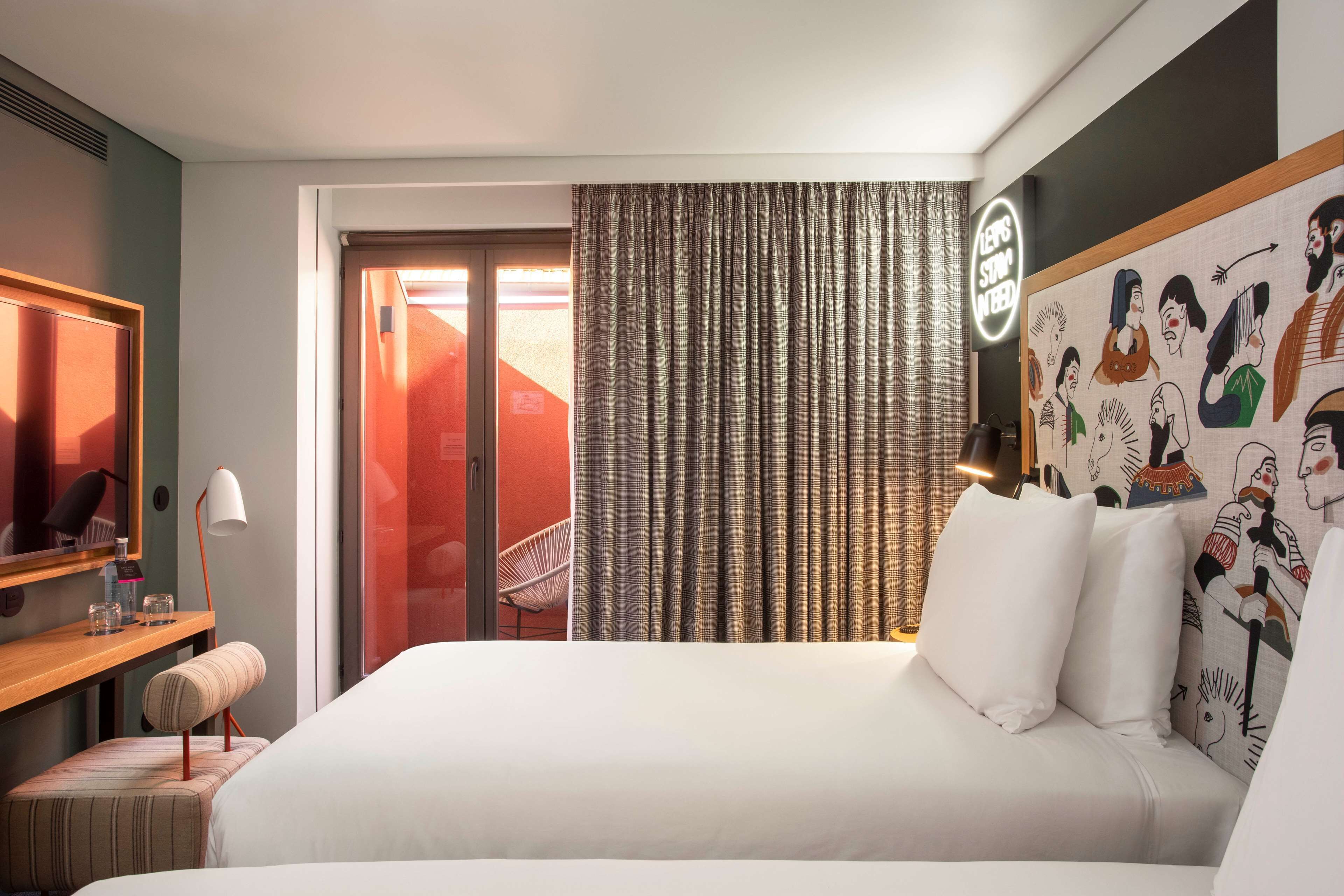 Ein Hotelzimmer mit Bett, Schreibtisch mit Lampe, Stuhl und einem großen Fenster mit Vorhängen.