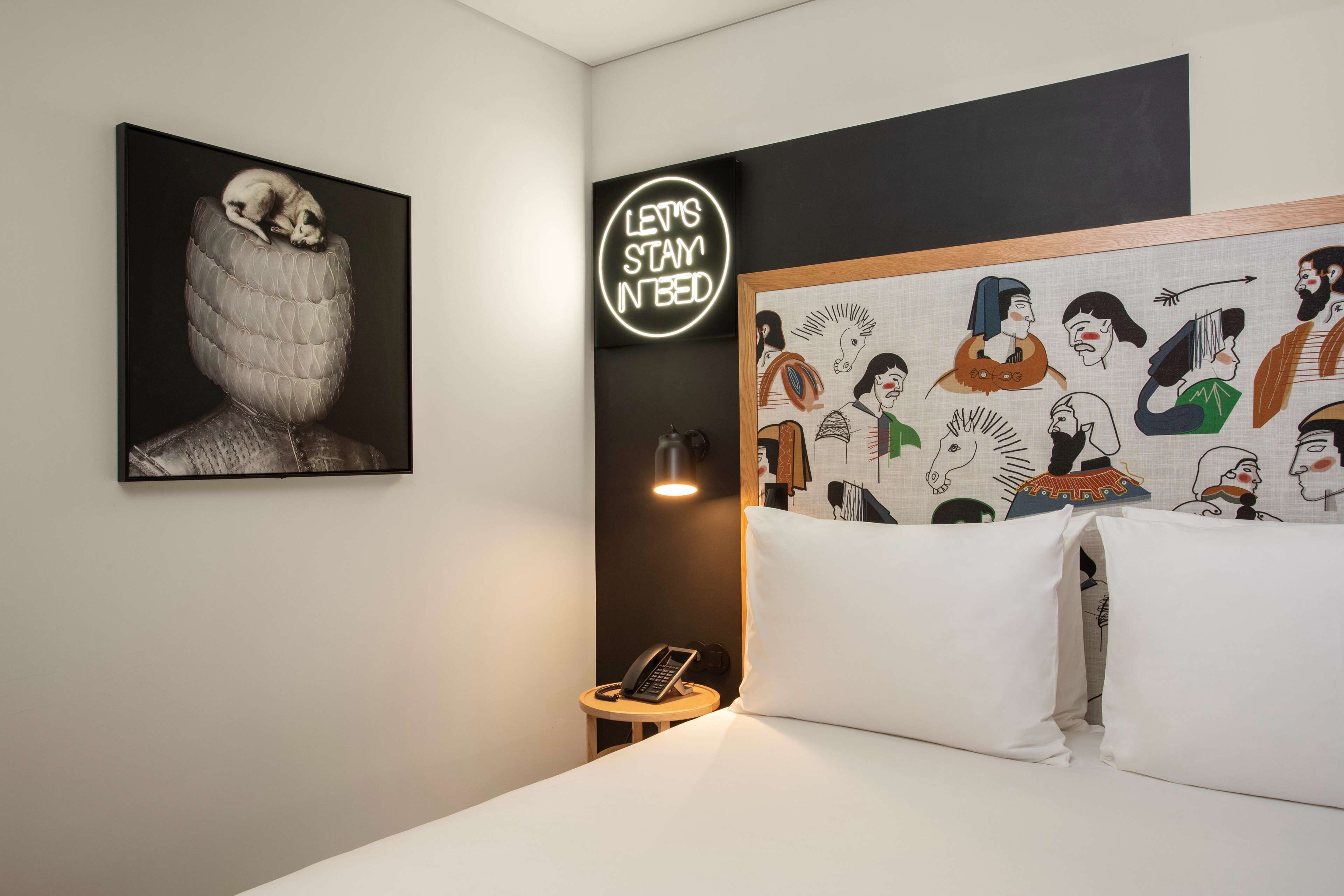 A hotel room with a bed, a bedside table with a phone, a wall-mounted light, and artwork including a neon sign and a mural.