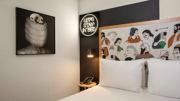 A hotel room with a bed, a bedside table with a phone, a wall-mounted light, and artwork including a neon sign and a mural.