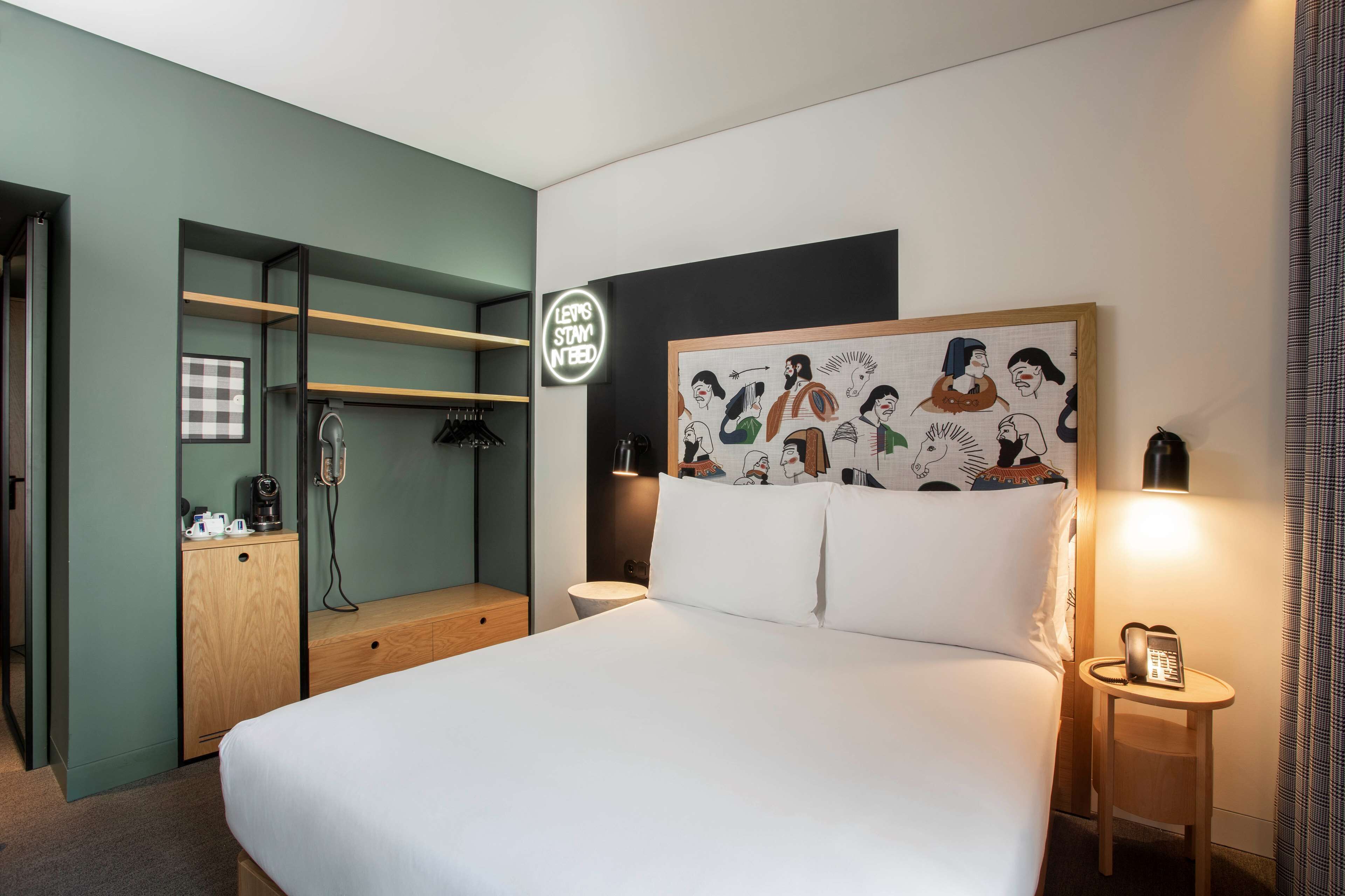 Quarto de hotel com uma cama, mesa de cabeceira com luminária, guarda-roupa e placa fixada na parede.