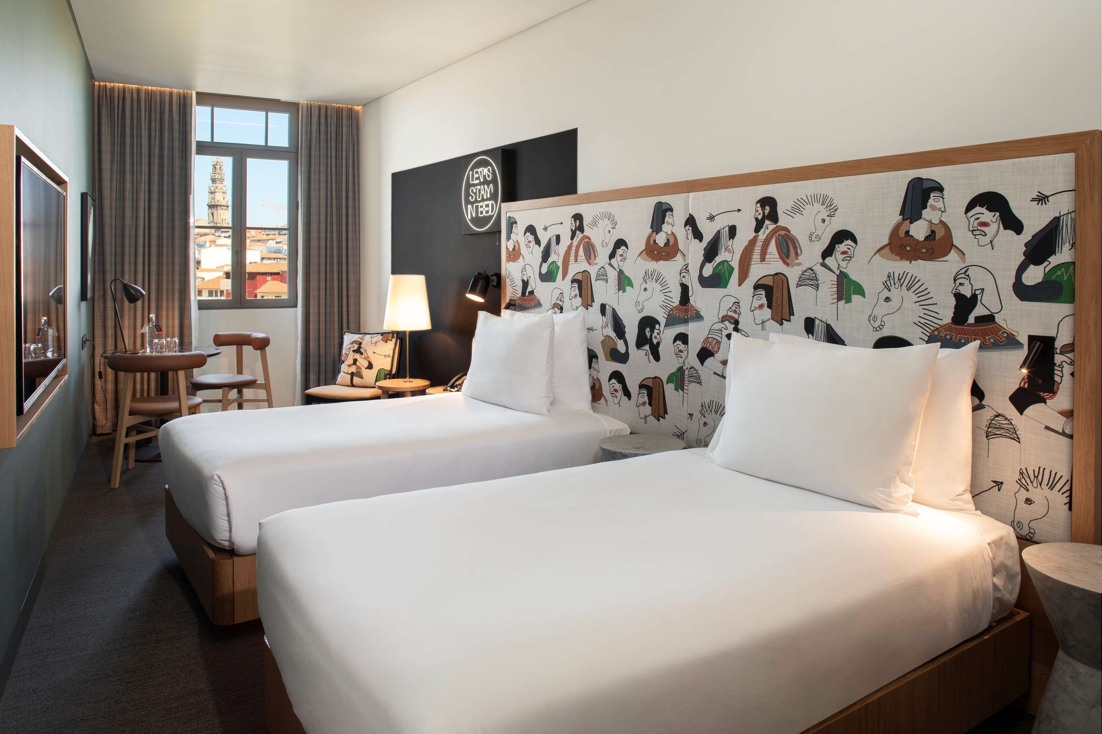 Een hotelkamer met twee bedden, een bureau, een stoel, een lamp en uitzicht op een stadsbeeld.