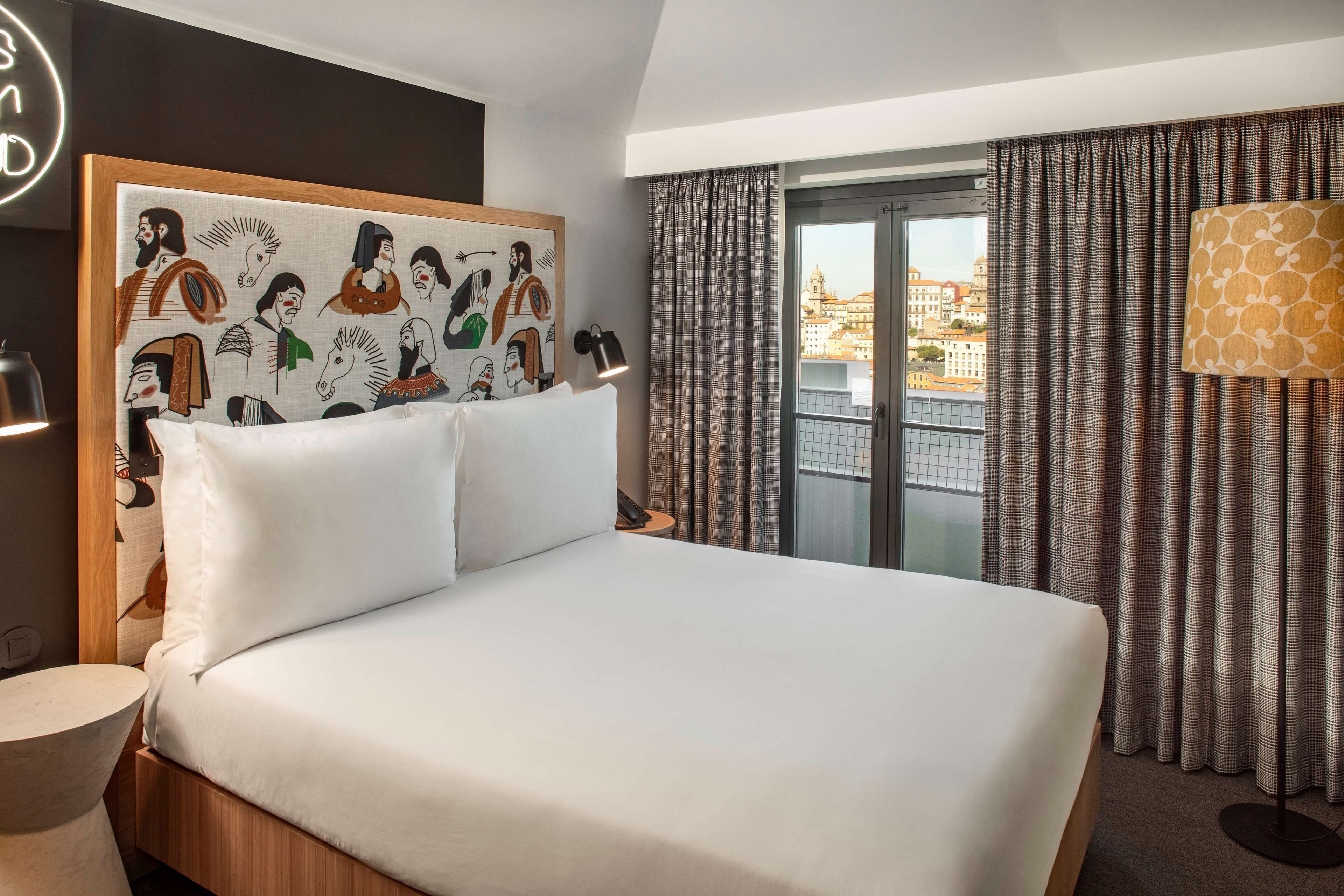 A hotel room with a large bed, a headboard with artwork, a view of the city through the window, and a lamp on the side table.