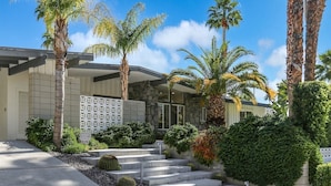 A modern building with a stone facade, a geometric white fence, and a well-maintained garden with palm trees and cacti. - Las Palmas Alexander – 1962 Alexander Home with Saltwater Pool, Spa, Fire Pit & Mountain Views (Palm Springs)