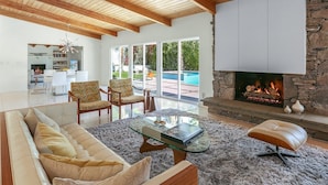 A modern living room with a stone fireplace, a glass coffee table, and a pool visible through the glass doors. - Las Palmas Alexander – 1962 Alexander Home with Saltwater Pool, Spa, Fire Pit & Mountain Views (Palm Springs)