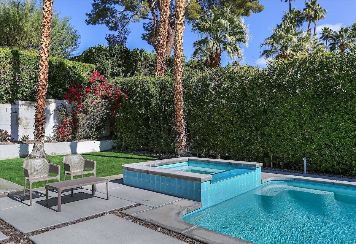 Une piscine dotée d’un banc intégré et de deux chaises, entourée d’un jardin bien entretenu avec des palmiers et des haies.