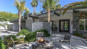 A modern outdoor patio with a fire pit, seating, and a stone wall. - Las Palmas Alexander – 1962 Alexander Home with Saltwater Pool, Spa, Fire Pit & Mountain Views (Palm Springs)