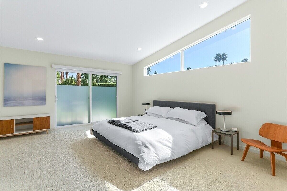 A modern bedroom with a large bed, a sliding glass door to a balcony, and a view of palm trees.