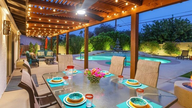 Un espacio para comer al aire libre con piscina, pérgola de madera y luces colgantes.