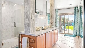 Un baño moderno con cabina de ducha de vidrio, zona de ducha con azulejos de mármol y tocador con lavamanos de cristal.