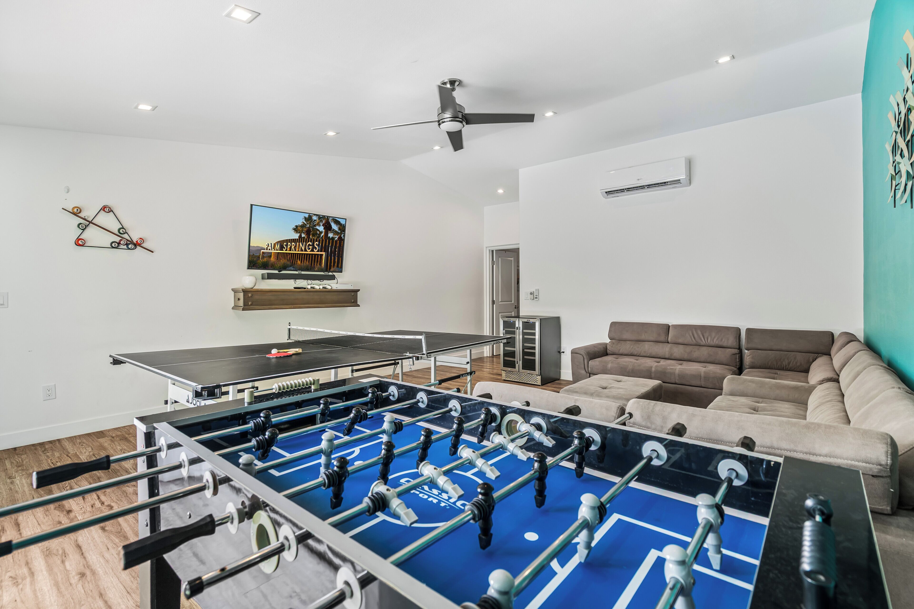 A room with a foosball table, a ping pong table, a sofa, and a television.