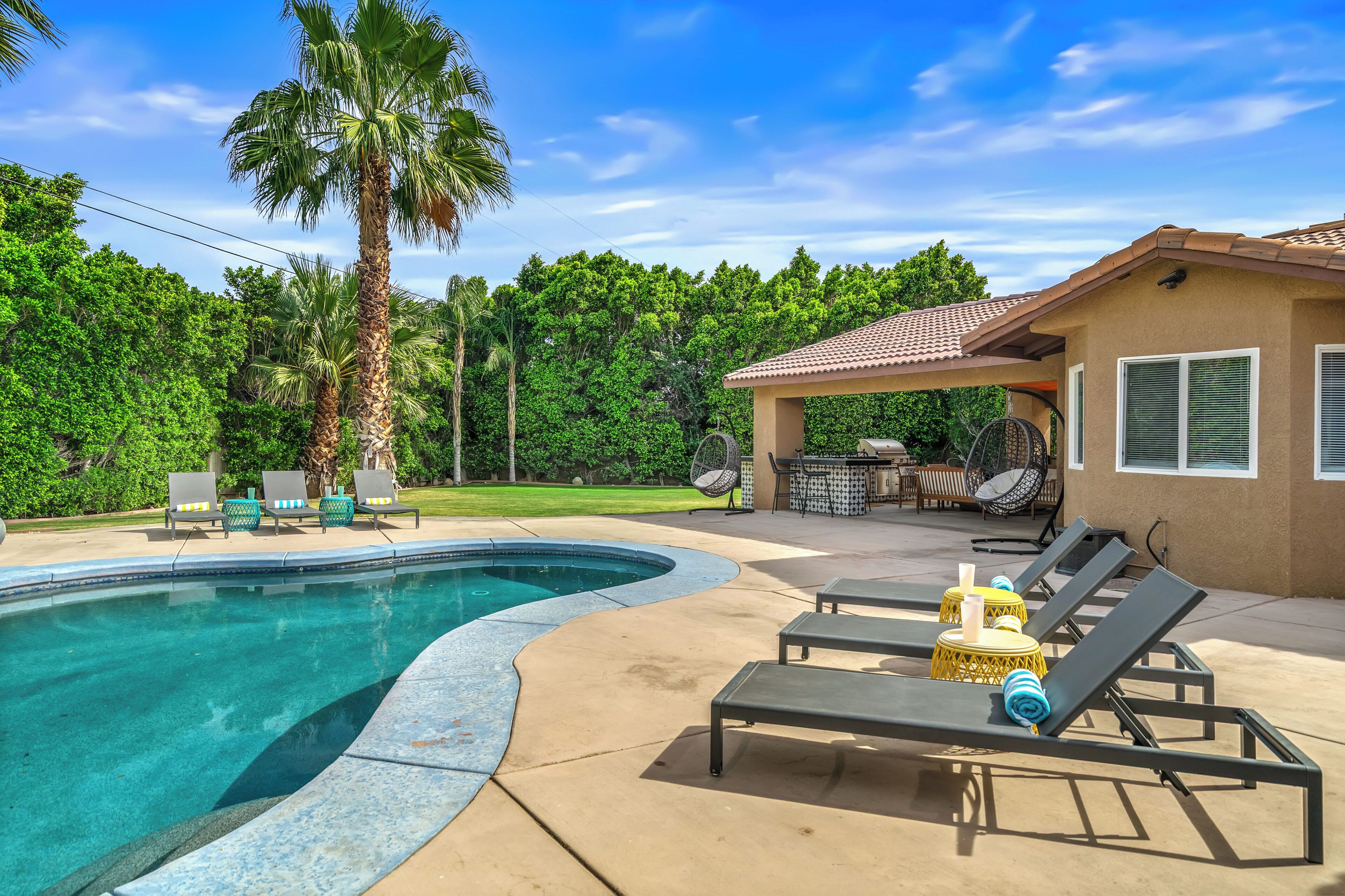 A swimming pool with lounge chairs, a covered patio, and a view of a lush green landscape.