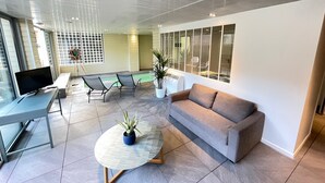 A modern living room with a grey sofa, a round coffee table with a plant, a TV on a stand, and a pool area visible through the glass wall. - Le Loft Mauresque Downtown Heated Indoor Pool (Arcachon)