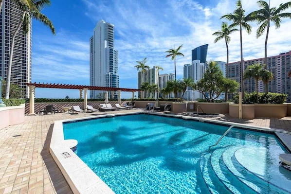 A swimming pool with lounge chairs and palm trees, surrounded by high-rise buildings.
