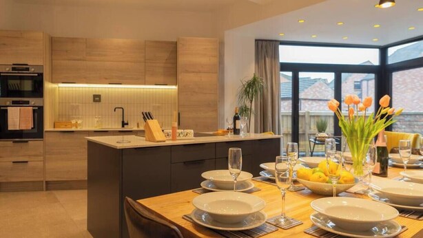 A modern kitchen with a central island, dining area with set tables, and large windows offering a view. - Escape 2 Unwind At The Parade, Accommodates 10 (York)