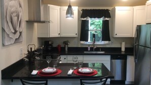 A modern kitchen with white cabinets, a black countertop, and a central island with red placemats. There is a window with black curtains, a stainless steel refrigerator, and a hanging pendant light. - Whidbey Island Orchard House Estate Langley, WA (Langley)