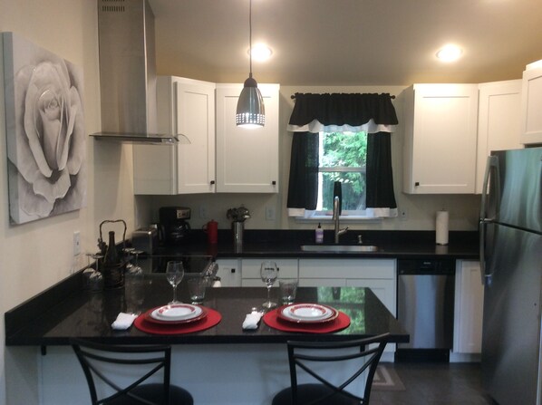 Una cocina moderna con armarios blancos, encimera negra y una isla central con individuales rojos. Hay una ventana con cortinas negras, un refrigerador de acero inoxidable y una lámpara colgante.