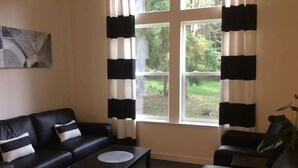 A living room with a black leather sofa, a coffee table, and a view of a garden through large windows with black and white curtains. - Whidbey Island Orchard House Estate Langley, WA (Langley)