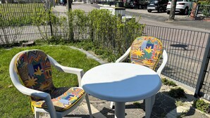 Ein kleiner Sitzbereich im Freien mit zwei weißen Kunststoffstühlen und einem weißen Tisch, umgeben von einem niedrigen Metallzaun und viel Grün.