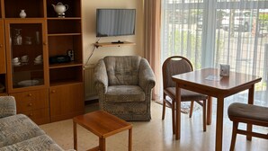 Ein Wohnzimmer mit Sofa, Sessel, Holzschrank, Fernseher und einem Esstisch mit Stühlen.