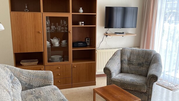Ein Wohnzimmer mit einer Couch, einem Couchtisch, einer Vitrine mit Gläsern, einem Fernseher und einem gemusterten Sessel.