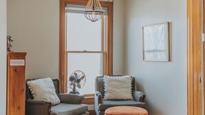 Une pièce avec un plancher en bois, deux fauteuils, un pouf rond, un ventilateur, un lustre, une fenêtre et une porte.
