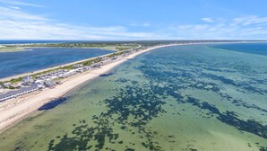 An aerial view of a coastal area with a long stretch of beach, clear water, and a row of buildings along the shore.